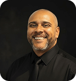 Michael Haynes smiling in a professional black shirt and tie, representing his role at Prosperia Marketing, showcasing the team's expertise and commitment to effective marketing strategies.