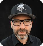 Travis Wachendorf, Prosperia Marketing team member, wearing a black cap and glasses, with a focused expression against a dark background.